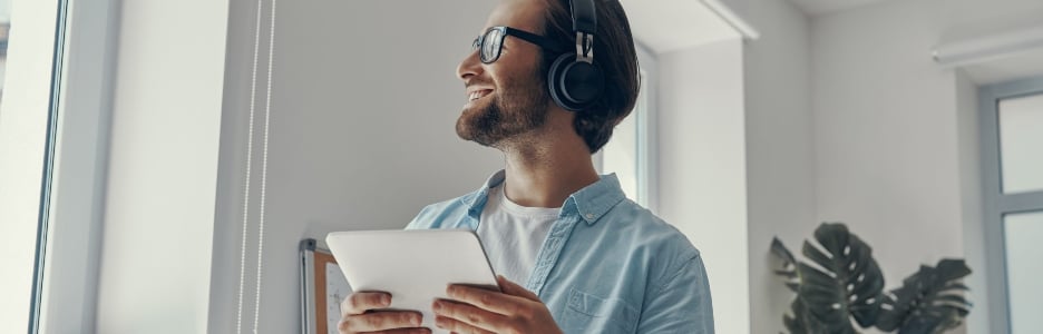 Imagen de un joven feliz con auriculares sosteniendo una tableta digital mientras está de pie en la oficina