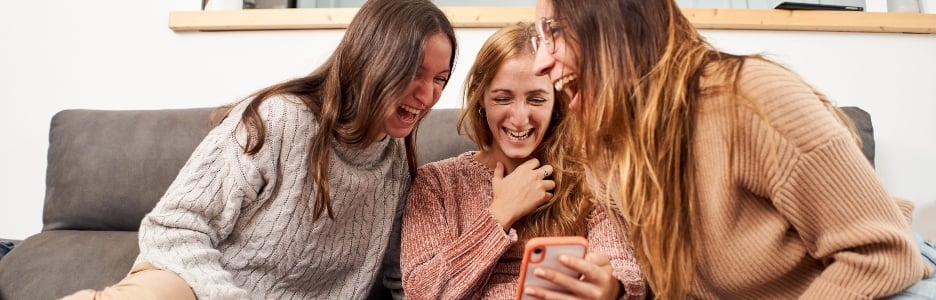Imagen de un grupo de amigas en el sofá de casa, usando un teléfono móvil riéndose