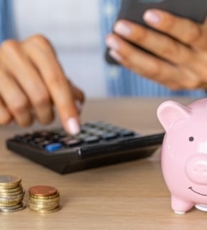 Una persona calculando sus finanzas con una calculadora y un móvil, junto a monedas apiladas y una hucha en forma de cerdito