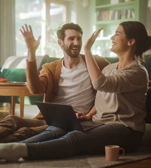 Imagen de una pareja usa una computadora portátil y choca las manos en señal de celebración mientras están sentados en el piso de la sala de su apartamento. Los novios conversan, se divierten, bromean, sonríen y se ríen.
