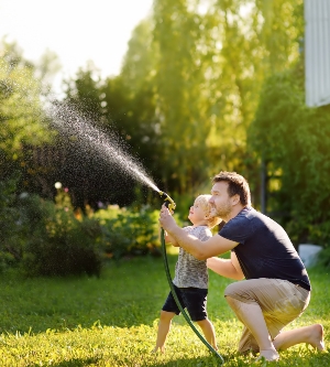 Imagen de un niño divertido con su padre jugando con una manguera de jardín en el soleado patio trasero. Niño en edad preescolar divirtiéndose con spray de agua. Actividad de verano al aire libre para niños.