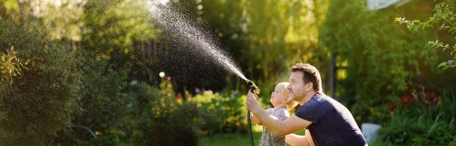 Imagen de un niño divertido con su padre jugando con una manguera de jardín en el soleado patio trasero. Niño en edad preescolar divirtiéndose con spray de agua. Actividad de verano al aire libre para niños.