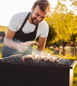 Imagen de un hombre alegre asando salchichas y carne en el campo