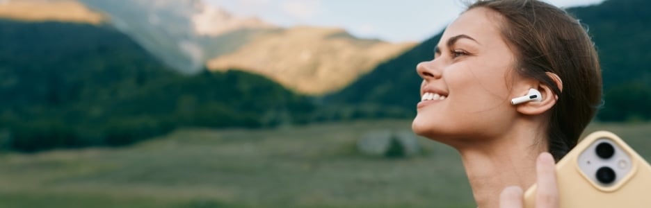 Imagen de una joven sonríe en un paisaje montañoso bañado por el sol mientras se toma un selfie informal con auriculares inalámbricos, disfrutando del aire fresco y el espacio abierto