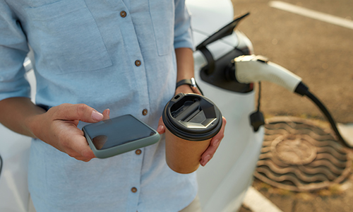 Imagen de una persona con café en mano cargando su coche eléctrico mientras navega con su teléfono móvil