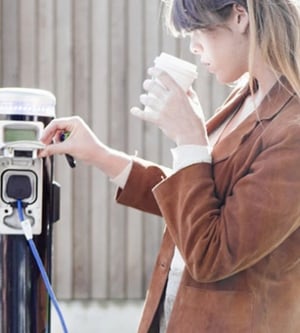 Imagen de una mujer cargando su coche eléctrico mientras se toma un café