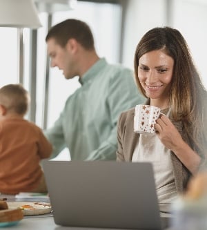 Imagen de una madre haciendo teleworking en su cocina mientras el padre de su hijo está jugando con el bebé