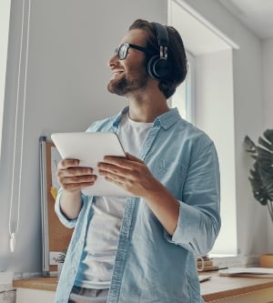 Imagen de un joven feliz con auriculares sosteniendo una tableta digital mientras está de pie en la oficina