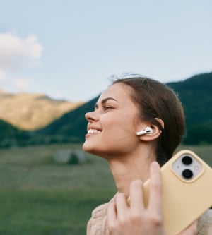Imagen de una joven sonríe en un paisaje montañoso bañado por el sol mientras se toma un selfie informal con auriculares inalámbricos, disfrutando del aire fresco y el espacio abierto