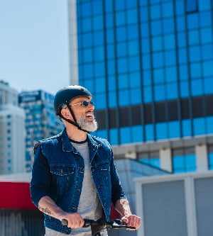 Imagen de hombre positivo y maduro con casco y gafas de sol conduciendo un patinete eléctrico en la calle de la ciudad
