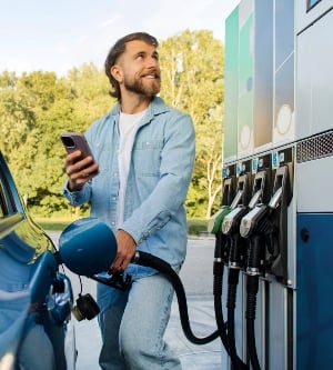Imagen de un hombre barbudo repostando el coche y usando el smartphone comparando los precios del combustible en la pantalla del surtidor y en el sitio web de la gasolinera