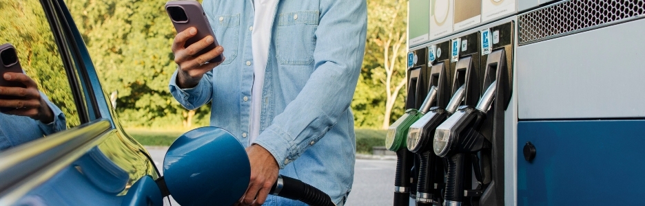 Imagen de un hombre barbudo repostando el coche y usando el smartphone comparando los precios del combustible en la pantalla del surtidor y en el sitio web de la gasolinera