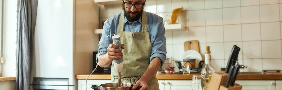 Imagen de joven, chef cocinero usando batidora de mano mientras prepara comida italiana en la cocina
