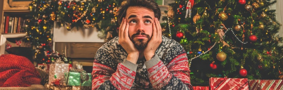 Imagen de un hombre joven sentado en un sofá rodeado de regalos bellamente envueltos y un árbol de Navidad decorado, reflexionando sobre los recuerdos a medida que se acerca el nuevo año